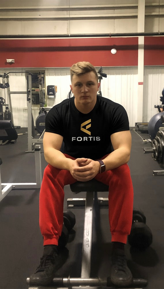 Founder of Fortis in gym wearing Fortis shirt, sitting on bench with dumbbells