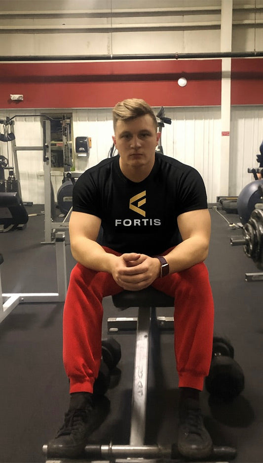 Founder of Fortis in gym wearing Fortis shirt, sitting on bench with dumbbells